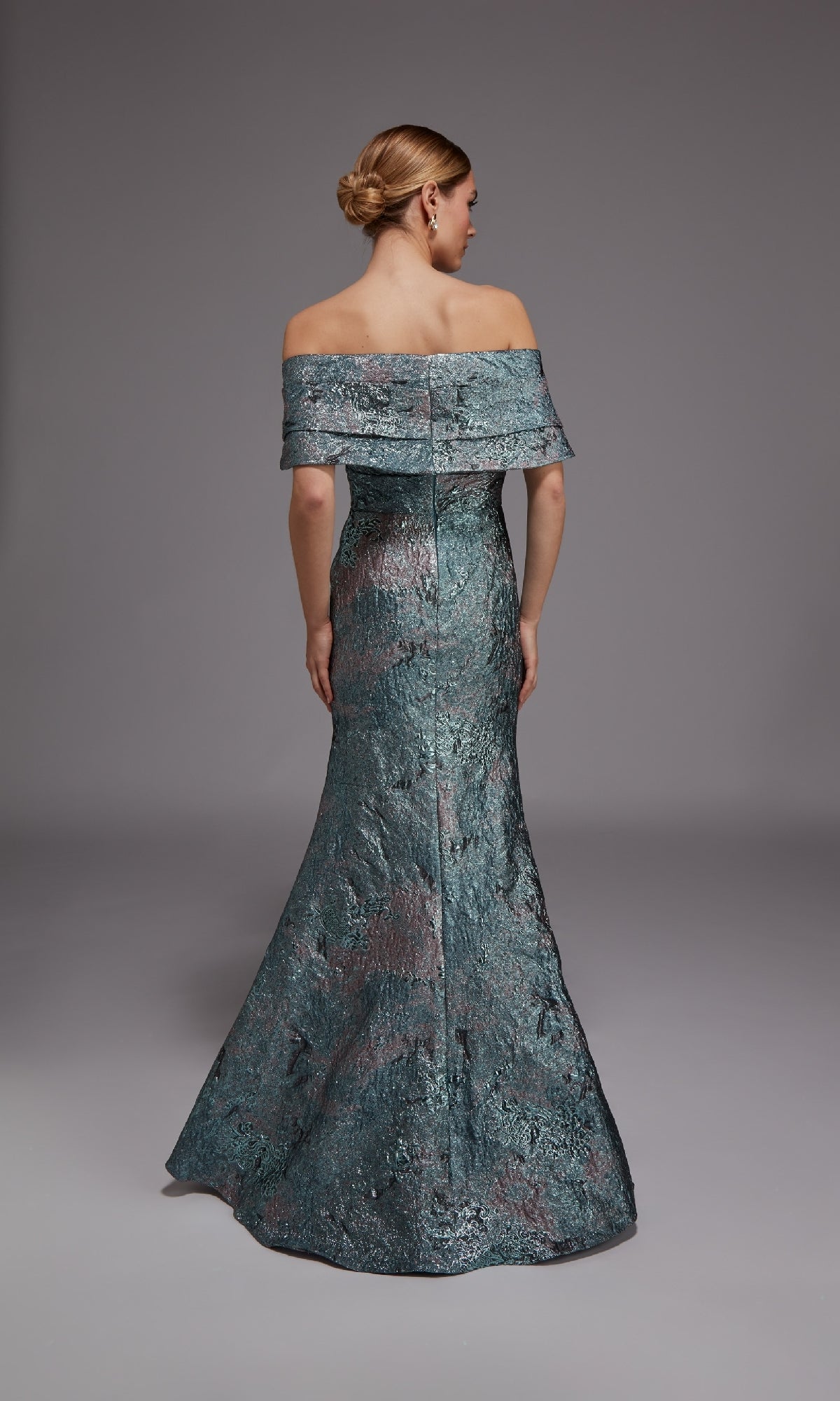 A woman faces away from the camera wearing the Alyce 27794, a metallic jacquard long formal dress featuring an off-the-shoulder neckline and flared hem. Her hair is styled in a sleek low bun, completing her elegant look for any formal occasion.